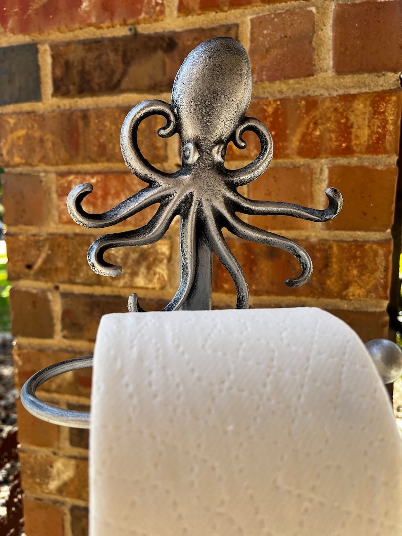 May include: Silver octopus-shaped toilet paper holder with a roll of white toilet paper. The holder is mounted on a brick wall.
