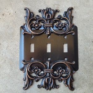 May include: A black and copper-toned decorative light switch cover with an ornate, scroll-like design. The cover has three toggle switches.