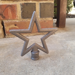 May include: A gray metal star-shaped decorative finial. The star has five points and is mounted on a cylindrical base. The finial is sitting on a concrete surface in front of a brick wall.