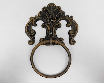 May include: A black and gold metal towel ring with a decorative, ornate design. The ring is attached to a wall mount with a scroll-like design.