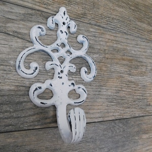May include: White cast iron wall hook with an ornate, scrolled design. The hook has a distressed, vintage appearance with black accents. Decorative wall hook for coats, hats, or other items.
