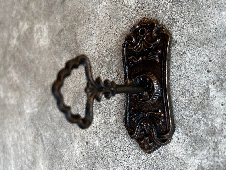Op de afbeelding: Omsiersleutel en sleutelplaat in antieke stijl met een bronzen afwerking. De sleutel is in het sleutelgat gestoken, wat toegang of veiligheid suggereert. De sleutelplaat heeft decoratieve bloemen- en kruldetails. De achtergrond is een getextureerd, grijs oppervlak.