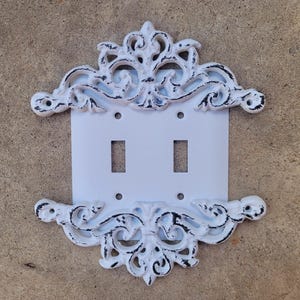 Pode incluir: Tampa de interruptor de luz dupla branca com uma moldura decorativa de ferro fundido branco adornada. A moldura tem um aspecto desgastado e envelhecido.