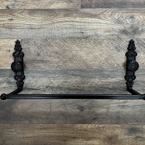 May include: Black metal towel bar with decorative scrollwork on the ends. The bar is mounted on a wood wall.