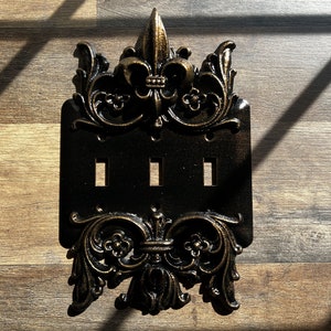 May include: A black and gold decorative light switch cover with a fleur-de-lis design. The cover has three switches.