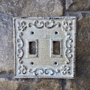 May include: Off-white double light switch cover with ornate scrollwork and floral accents. The cover has two rectangular openings for the switches and four screw holes. The edges are distressed with dark brown.