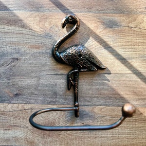 Puede incluir: Un porta rollo de papel higiénico de metal negro en forma de flamenco con una base redonda. El soporte está montado en una pared de madera.