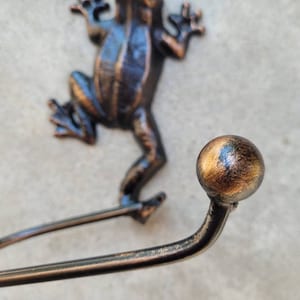 May include: A cast iron frog wall hook with a decorative gold-toned ball finial. The hook is designed to hold towels, robes, or other items.
