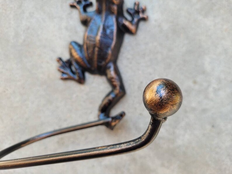 May include: A cast iron frog wall hook with a decorative gold-toned ball finial. The hook is designed to hold towels, robes, or other items.