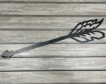 May include: Black metal arrow with a decorative feather design. The arrow is about 24 inches long and is mounted on a light gray wood background.