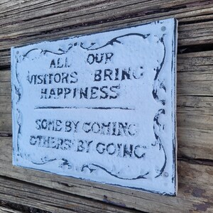 May include: White sign with black lettering that reads "All our visitors bring happiness, some by coming, others by going". The sign has a decorative border and is mounted on a wooden surface.