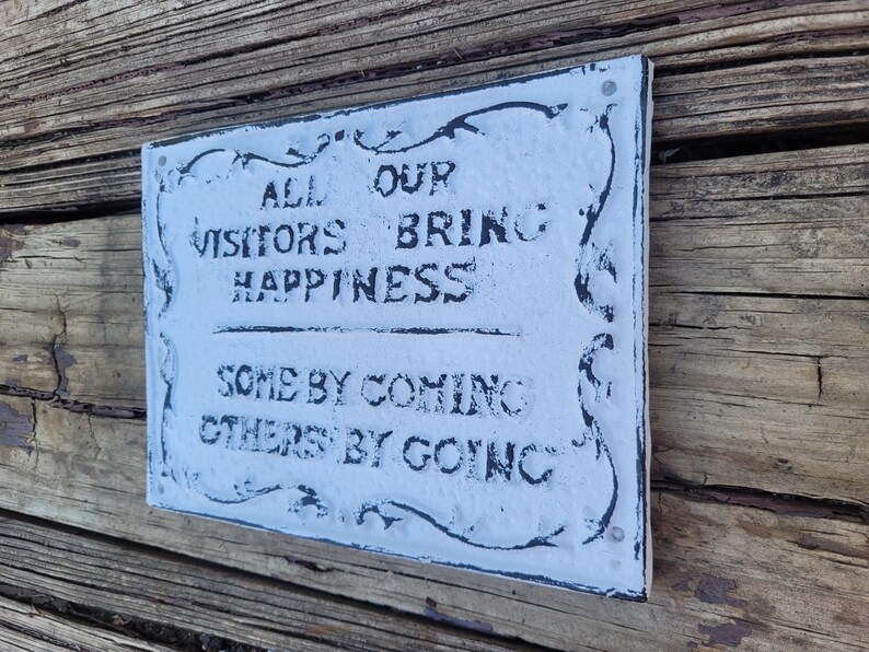 May include: White sign with black lettering that reads "All our visitors bring happiness, some by coming, others by going". The sign has a decorative border and is mounted on a wooden surface.