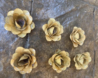 Puede incluir: Seis adornos de flores de metal hechos a mano en varios tamaños. Las flores están pintadas en un amarillo apagado con centros más oscuros, dispuestas sobre una superficie texturizada similar a la piedra. Estas flores decorativas podrían usarse para manualidades o decoración del hogar.