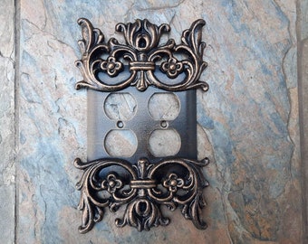May include: A decorative outlet cover with an ornate, bronze-colored metal design. The cover has two outlets and is designed to fit a standard electrical outlet.
