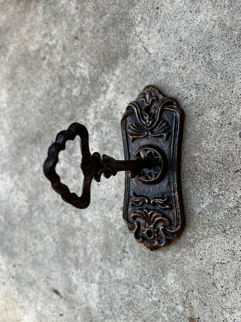Op de afbeelding: Ouderwetse sleutel en sleutelplaat in antieke stijl. De sleutel heeft een decoratieve, cirkelvormige handgreep en een gedetailleerd, bloemachtig ontwerp. De rechthoekige sleutelplaat heeft een uitgebreid, symmetrisch patroon. Het metaal heeft een donkere, verouderde afwerking.