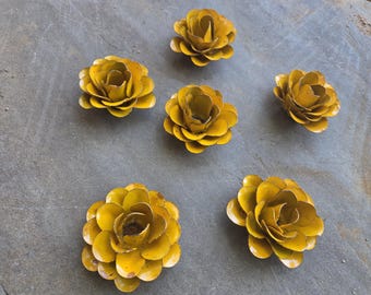 Puede incluir: Seis decoraciones florales de metal amarillo con pétalos en capas están dispuestas sobre una superficie gris. Las flores tienen un aspecto ligeramente desgastado, lo que sugiere que pueden ser para la decoración del jardín o del hogar.