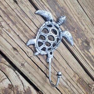 May include: Silver metal turtle hook with a detailed shell design. The hook is mounted on a wooden surface.
