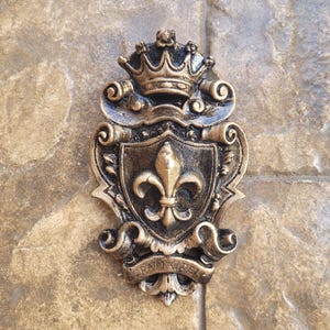 May include: A gold-colored metal coat of arms with a crown, a fleur-de-lis, and a scroll with the text "GRATI A DEI".