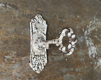Puede incluir: Un gancho de pared con una placa decorativa ornamentada y un brazo que sobresale con forma de llave antigua. El objeto de metal tiene un acabado blanco desgastado y envejecido con acentos marrón oscuro en la superficie texturizada.