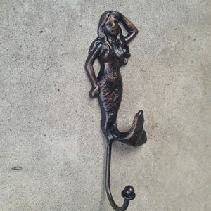 May include: A cast iron mermaid hook with a dark brown finish. The mermaid is depicted with long flowing hair and a tail that forms the hook.
