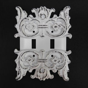 Pode incluir: Tampa de interruptor de luz decorativa branca com um design ornamentado e desgastado. A tampa é feita de metal fundido e tem um interruptor duplo.