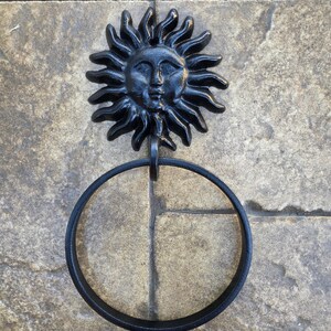 May include: A black metal sun face towel ring. The sun has a detailed face and radiating rays. A circular ring hangs below the sun, designed to hold a towel. The fixture is mounted on a stone tile surface.