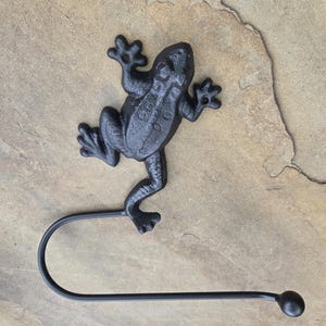 May include: Black cast iron frog-shaped paper towel holder. The frog is detailed with textured skin and outstretched limbs. The holder has a curved arm with a ball-shaped end. A decorative and functional item.