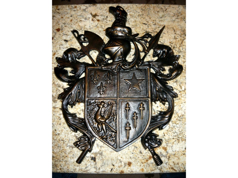 May include: A bronze metal coat of arms with a shield, helmet, and other heraldic symbols. The shield is divided into four sections, each with a different design. The top left section features three fleurs-de-lis, the top right section features a five-pointed star, the bottom left section features a bird, and the bottom right section features five crosses.