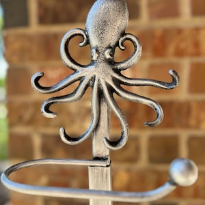 May include: Silver metal octopus toilet paper holder. The octopus has a detailed design and is mounted on a silver metal pole. The holder has a round base.