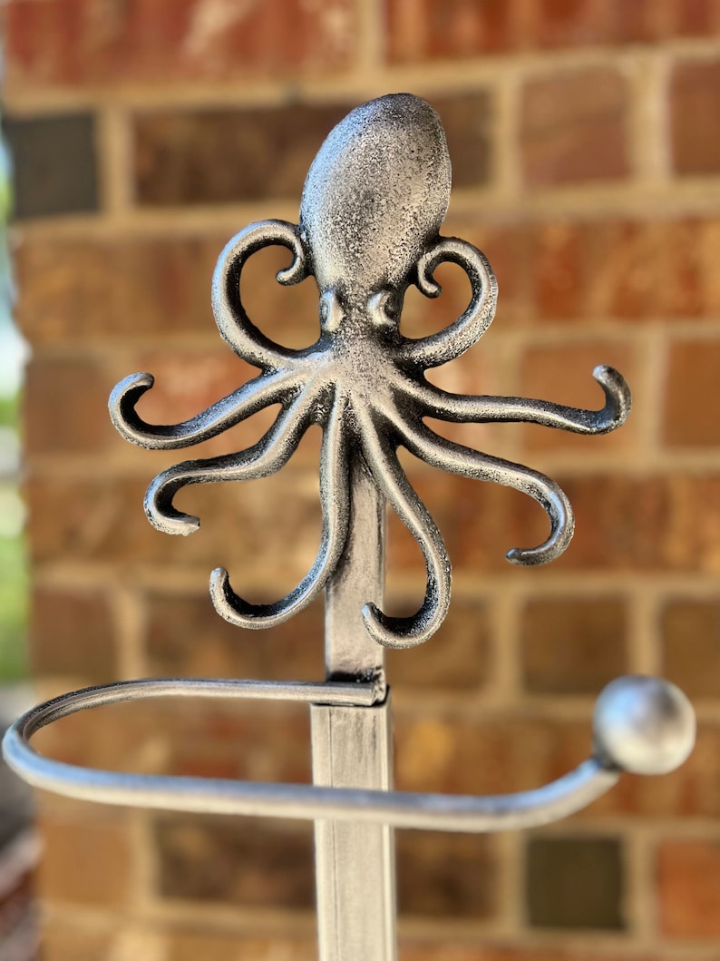 May include: Silver metal octopus toilet paper holder. The octopus has a detailed design and is mounted on a silver metal pole. The holder has a round base.