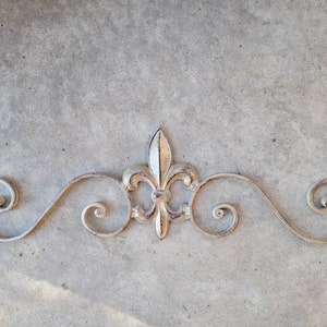 May include: A decorative metal wall decor featuring a fleur-de-lis design with a white and gold finish. The design is made of a series of swirls and curves.