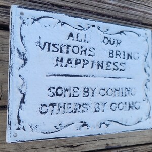 May include: White metal sign with black lettering that reads "All our visitors bring happiness, some by coming, others by going."