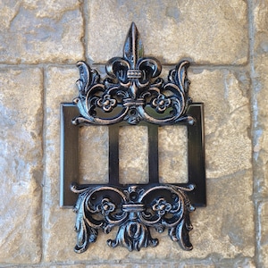 Könnte beinhalten: Ein schwarzer dreifacher Lichtschalter aus Metall mit einem verzierten Fleur-de-lis-Design.
