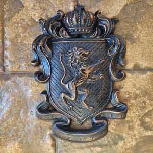 Op de afbeelding: Een zwart metalen wapenschild met een leeuw die opstaat en een kroon bovenop. Het wapenschild is versierd met rollen en heeft een gouden afwerking.