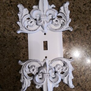 May include: White decorative light switch cover with a fleur-de-lis design. The cover is made of metal and has a distressed finish.