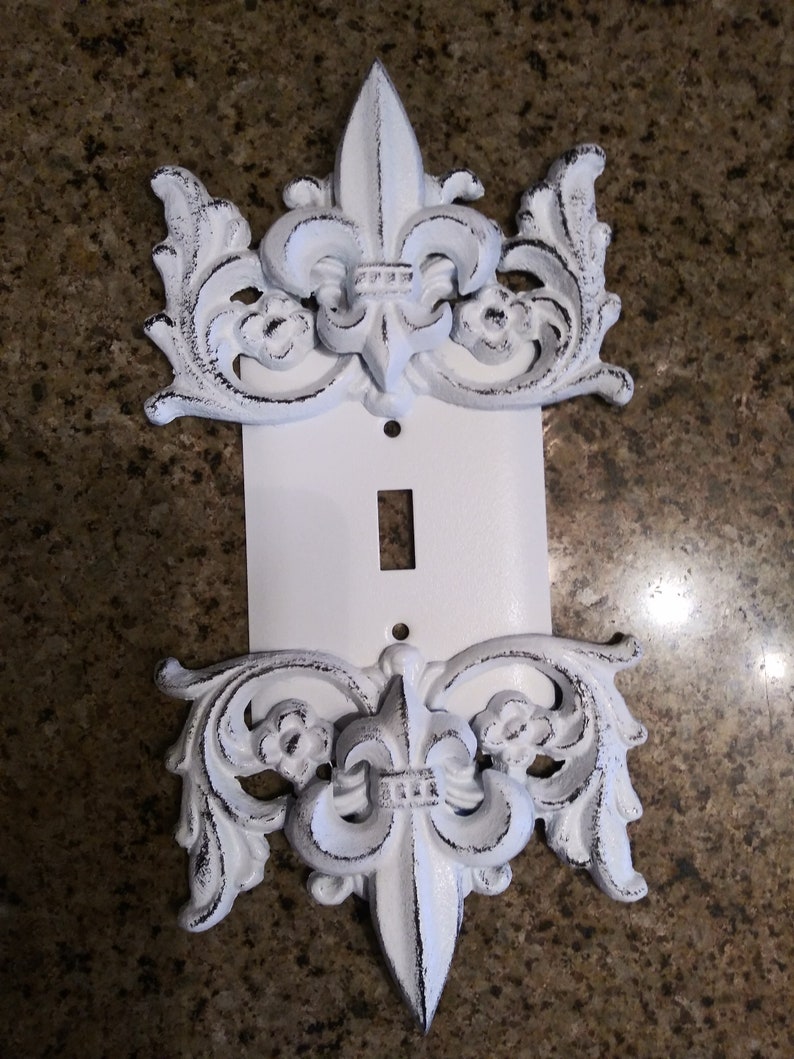 May include: White decorative light switch cover with a fleur-de-lis design. The cover is made of metal and has a distressed finish.