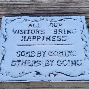 May include: A white sign with black lettering that reads "All our visitors bring happiness, some by coming, others by going". The sign has a decorative black border.