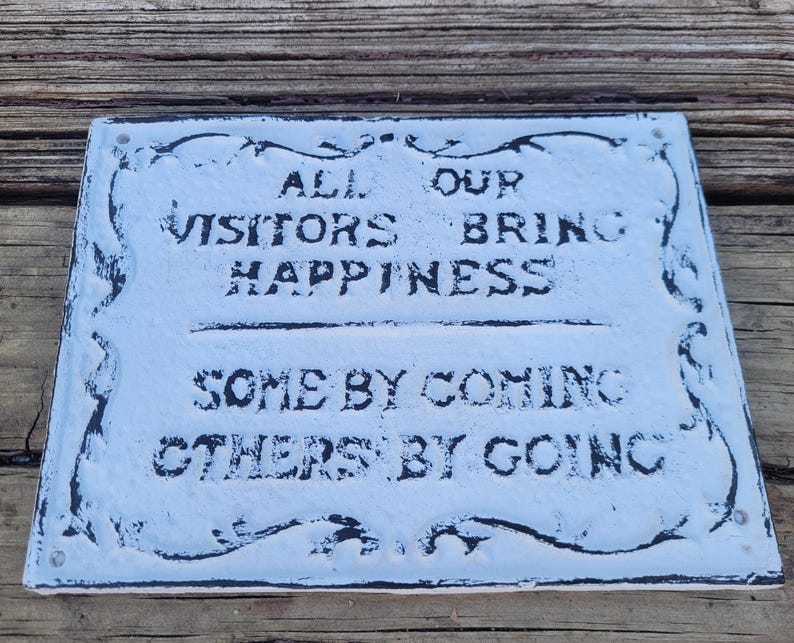 May include: A white sign with black lettering that reads "All our visitors bring happiness, some by coming, others by going". The sign has a decorative black border.