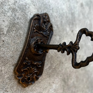 Op de afbeelding: Een antieke bronzen sleutel die in een bijpassende sleutelplaat is gestoken. De sleutel heeft een sierlijke handgreep en de plaat heeft decoratieve bloemen- en kruldetails. De sleutel en de plaat staan tegen een getextureerde, grijze achtergrond.