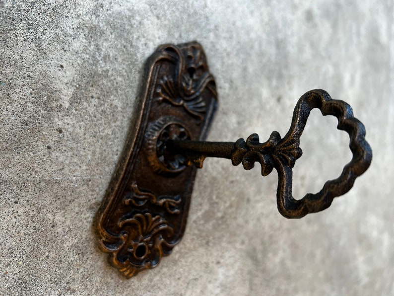 Op de afbeelding: Een antieke bronzen sleutel die in een bijpassende sleutelplaat is gestoken. De sleutel heeft een sierlijke handgreep en de plaat heeft decoratieve bloemen- en kruldetails. De sleutel en de plaat staan tegen een getextureerde, grijze achtergrond.