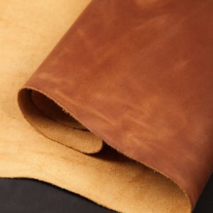 Op de afbeelding: Close-up van twee stukken leer, één opgerold en de andere plat. Het opgerolde leer is een rijk, warm bruin, terwijl het platte stuk een lichtere, bruine kleur heeft. De texturen van het leer zijn zichtbaar, wat wijst op een natuurlijk materiaal.