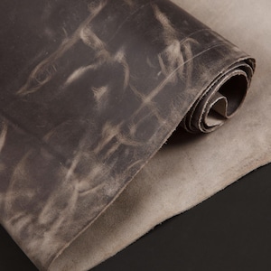May include: A rolled-up piece of distressed leather in shades of gray and brown. The leather has a textured surface with lighter markings, and the edges are visible. The background is a solid black.
