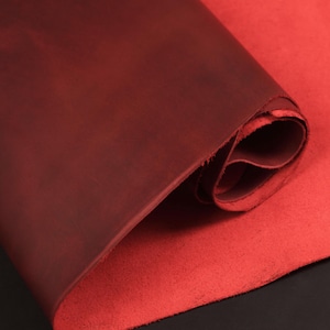 May include: A rolled-up piece of red leather, with a darker shade of red on the top layer. The edges of the leather are visible, showing the texture and thickness of the material. The leather appears to be soft and pliable.