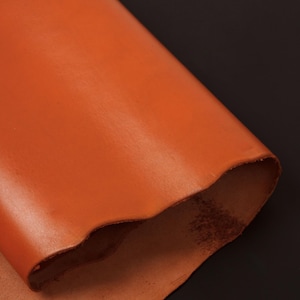 May include: A close-up of a piece of orange leather. The leather is smooth and has a slight sheen. It is folded in half and the edges are slightly curled.