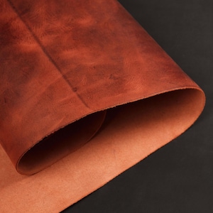 May include: Close-up of a rolled sheet of reddish-brown leather. The leather has a textured surface with variations in color, and the edges are visible, revealing a lighter shade of brown. The background is black.