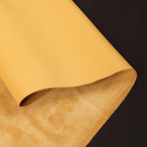 May include: A sheet of yellow leather, folded over to reveal a textured surface and a softer, suede-like underside. The leather has a smooth, matte finish and is set against a dark background.