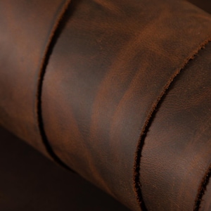 May include: Close-up of a rolled piece of brown leather. The leather has a distressed appearance with variations in color, ranging from dark brown to lighter shades. The edges of the leather are visible, showing a raw, unfinished texture.