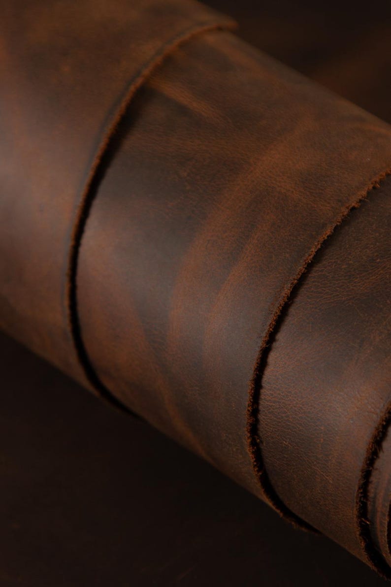 May include: Close-up of a rolled piece of brown leather. The leather has a distressed appearance with variations in color, ranging from dark brown to lighter shades. The edges of the leather are visible, showing a raw, unfinished texture.