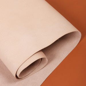 Puede incluir: Una hoja enrollada de cuero beige claro, que muestra una superficie texturizada. El cuero está parcialmente desenrollado, revelando una parte inferior lisa. El fondo es de color naranja cálido sólido. Esta imagen resalta la textura y el color del cuero.