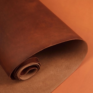 May include: A rolled-up piece of brown leather, showcasing a rich, textured surface. The leather has a warm, reddish-brown hue, with visible grain and subtle variations in color. The edges of the roll reveal a lighter, tan underside.